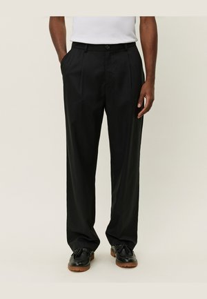 Homme portant un pantalon noir ample sur mesure avec des poches latérales, associé à des mocassins noirs à glands et une chemise blanche rentrée.
