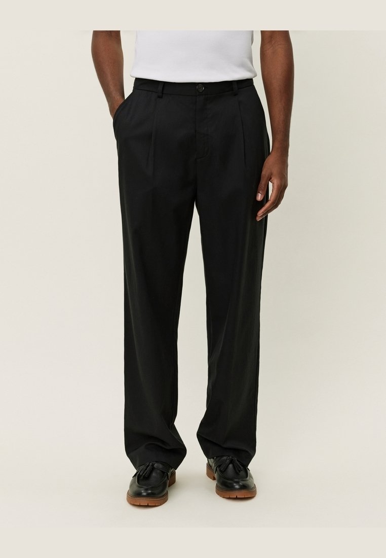 Homme portant un pantalon noir ample sur mesure avec des poches latérales, associé à des mocassins noirs à glands et une chemise blanche rentrée.