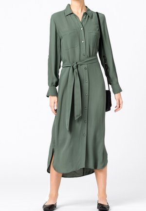 Mujer con vestido midi de manga larga color verde oliva, botones delanteros, bolsillos frontales, cinturón de tela, aberturas laterales, zapatos planos negros y bolso negro de hombro.