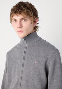 Gray zip-up sweater with a high collar, ribbed texture, and a small embroidered logo on the left chest. Made from soft knit material.