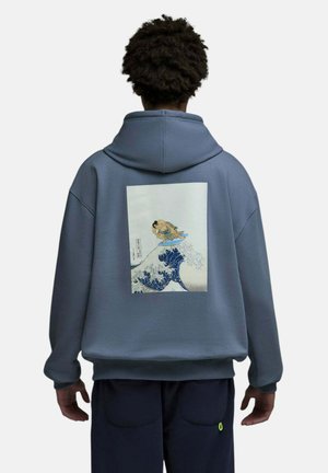 Personne portant un sweat à capuche bleu avec un motif de style japonais représentant un lutteur de sumo chevauchant une vague dans le dos, debout devant un fond blanc.