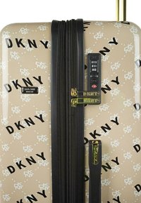 Maleta rígida en beige con patrón de logotipos en negro de DKNY, que cuenta con un candado de combinación, cremalleras negras y detalles dorados en el asa.