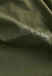 Olivenfarget bomullsstoff har en subtil tekstur med en stor, mørkegrønn logo med teksten "JACK & JONES" trykket over overflaten.
