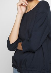 Eine Person trägt ein lockeres, marineblaues Langarmshirt mit teilweise hochgerollten Ärmeln und hat die Hand nahe dem Kinn auf einem schlichten Hintergrund abgelegt.