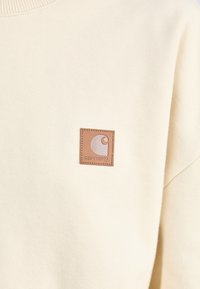 Sweatshirt crème avec un col ribbé, doté d'un logo en patch carré en cuir marron sur la poitrine gauche. Texture douce et design décontracté.