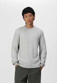 Un pull en tricot côtelé gris clair avec un col rond et des manches longues, présentant un tissu texturé et un ourlet ajusté, porté sur un pantalon sombre.