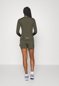 Frau mit langen geflochtenen Haaren, die ein olivgrünes Langarmoberteil und Shorts trägt, weiße Socken und graue Sportschuhe, steht mit dem Rücken zu uns.