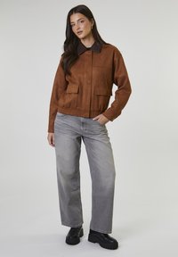 Kobieta stojąca z jedną ręką w kieszeni, ubrana w brązową kurtkę z klapkami, szare szerokie dżinsy i czarne buty na platformie na jednolitym tle.