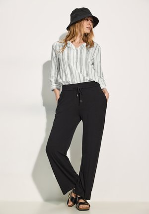 Camisa de botones gris claro con rayas verticales negras, combinada con pantalones negros de corte holgado y sandalias negras. El modelo lleva un sombrero de cubo negro.