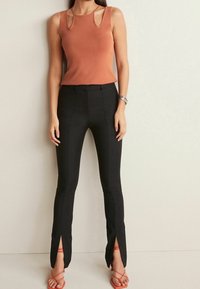 Rustkleurig mouwloos top met uitgesneden schouderdetails, gecombineerd met hooggetailleerde zwarte broek met zijslits en rode sandalen met banden.