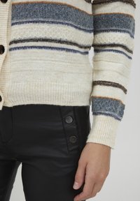 Adīta kardigāna ar horizontālām svītrām krēmkrāsā, zilā, brūnā un pelēkā krāsā. Iezīmējas ar teksturētu audumu un melnām pogām uz piedurknēm.