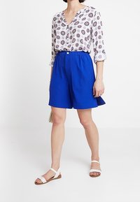 Weiße Bluse mit einem kreisförmigen roten und marineblauen Muster, kombiniert mit blauen Shorts mit elastischem Bund und Seitentaschen. Getragen mit weißen Sandalen.