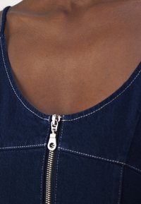 Jeans oblačilo s krojaškim izrezom, ki predstavlja vidno srebrno zadrgo in kontrastno belo šivanje na temno modri tkanini. Gladka tekstura.