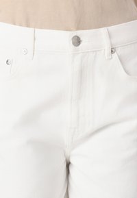 Los jeans de mezclilla blanca presentan un diseño clásico de cinco bolsillos, cierre con botón plateado y una tela texturizada con sutiles patrones de tejido diagonal.