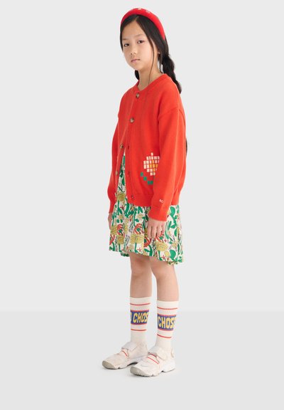 Niña joven vistiendo un cárdigan rojo, vestido floral, boina roja, calcetines blancos con texto azul y amarillo, y zapatillas blancas, de pie frente a un fondo gris.