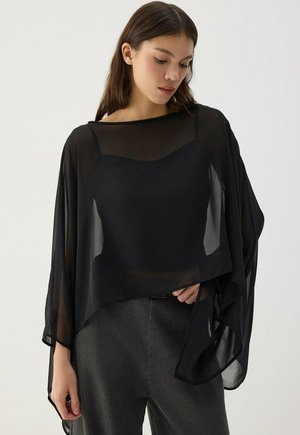 Jeune femme aux longs cheveux bruns porte un haut noir transparent et ample par-dessus un caraco noir et un pantalon gris foncé, regardant légèrement vers le bas.