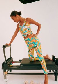 Set sportiv colorat, format dintr-un top scurt și colanți cu modele tropicale. Modelul pozează pe o mașină de reformare neagră.