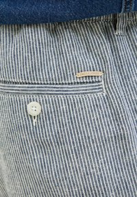 Dryžuota mėlyna ir balta medžiaga su mygtukų uždarymu; turi mažą odinę etiketę su užrašu "JACK & JONES" šalia kišenės viršaus.