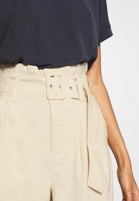 Närbild av en person som bär beige högmidjade byxor med ett brett bälte och rektangulär spänne, ihop med en mörk marinblå t-shirt som är stoppad i byxorna.