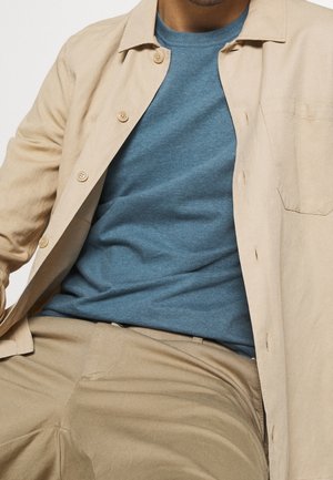 Mann trägt beige Hemd über blauem Rundhals-Pullover und beigen Hosen, sitzt vor einem einfarbigen Hintergrund.