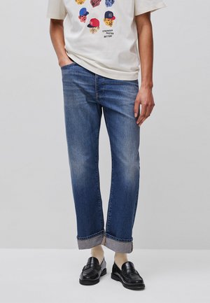 Person mit hochgekrempelten blauen Jeans, cremefarbenen Socken, schwarzen Loafers und einem weißen T-Shirt mit Comic-Gesichtern und dem Text „Stronger Faster Better!“.