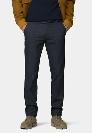 Mann trägt dunkelblaue Slim-Fit-Hose, braunen Gürtel, braune Schnürstiefel, marineblauen Pullover und senffarbene Jacke mit den Händen in den Taschen.