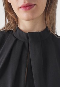 Black blouse featuring a high neckline with a folded detail and pleated front, made from a lightweight, smooth fabric.