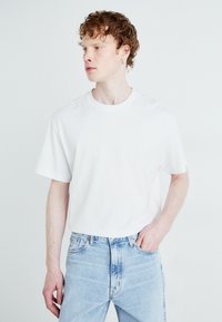 Fehér pamut póló, rövid ujjakkal és kerek nyakkivágással, világoskék farmernadrággal párosítva. Simaság, laza szabás.