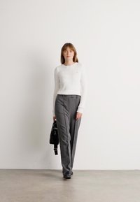 Vrouw die binnen loopt in een witte kabelgebreide trui, grijze hoge taillebroek, zwarte schoenen en een zwarte handtas draagt.