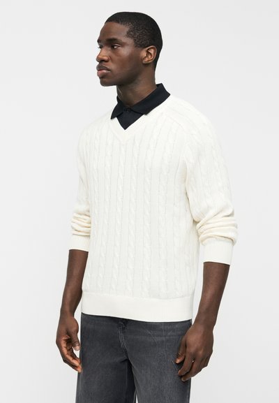 Άνδρας που φοράει λευκό πουλόβερ με κόμπους και V-λαιμόκοψη πάνω από μαύρο πουκάμισο με γιακά, συνδυασμένο με σκούρα γκρι τζιν, στέκεται μπροστά σε απλό φόντο.