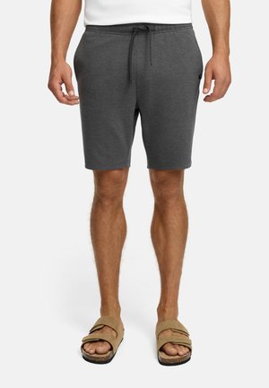 Mand i mørkegrå snøre shorts og beige åbent tå-sandaler, der står op ad en hvid baggrund.