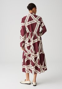 Abito bordeaux con motivi astratti bianchi, maniche lunghe, design a strati e colletto. Indossato con mocassini di colore chiaro, vista posteriore.