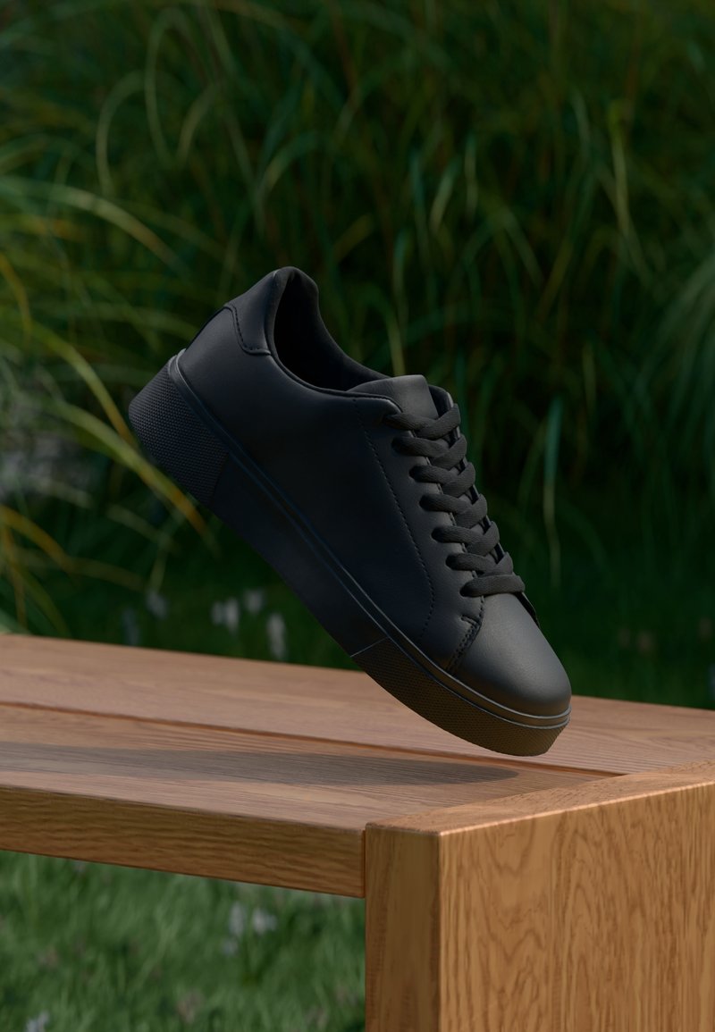 Baskets noires à lacets flottant au-dessus d'un banc en bois, installées en extérieur avec une haute herbe verte en arrière-plan.