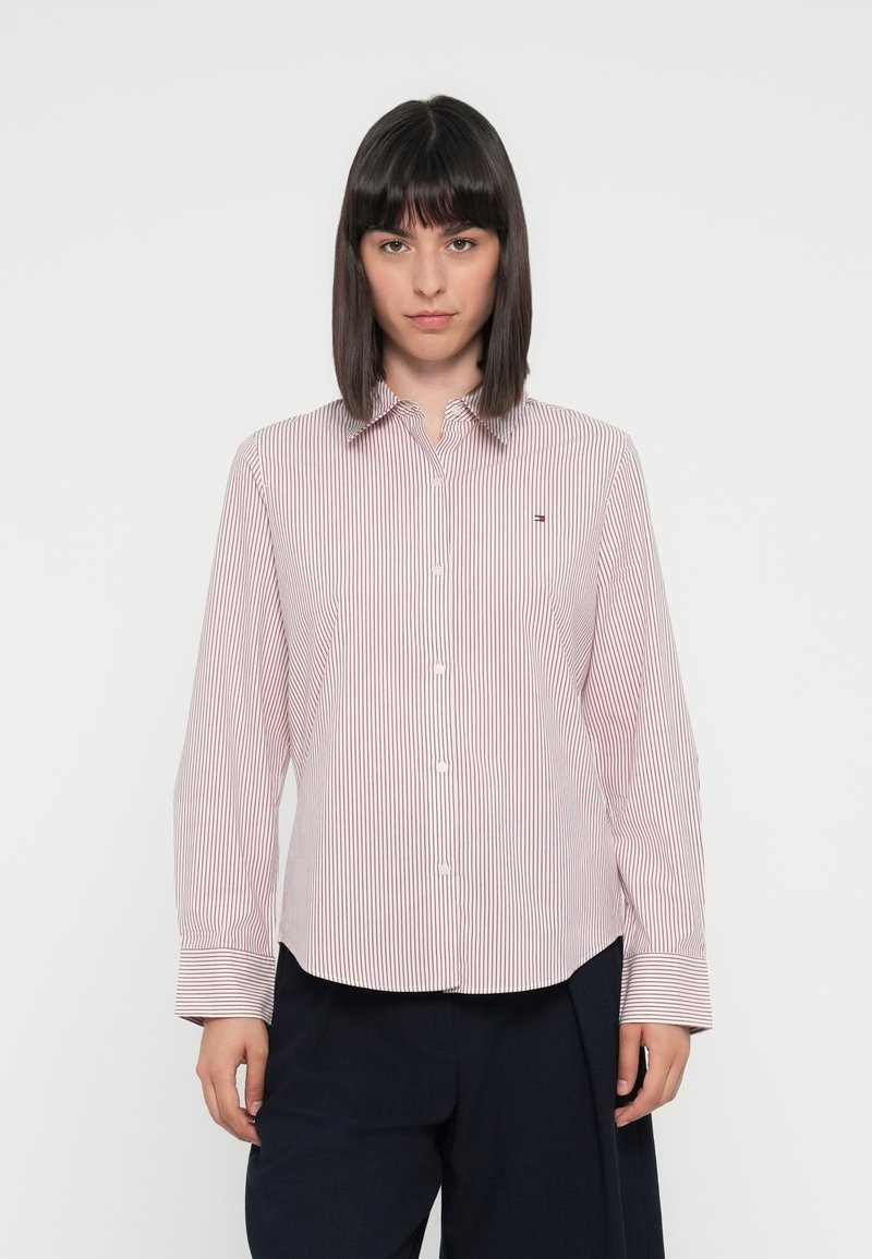 Chemise boutonnée à rayures rouges et blanches avec un col classique, des manches longues et un petit logo sur la poitrine. Texture de tissu lisse.