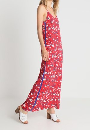 Femme portant une robe maxi rouge sans manches avec un imprimé floral blanc et des bandes latérales bleues, associée à des sandales blanches à talons.