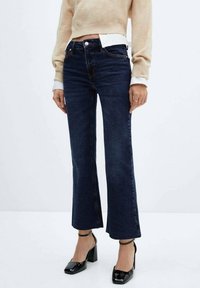 Jean évasé bleu foncé en denim, avec une taille haute et un ourlet brut, associé à des chaussures noires à talons blocs et un pull beige.