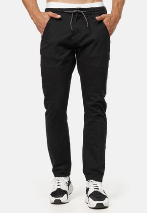 Man draagt zwarte joggerbroek met een tunnelkoord in de taille en witte en zwarte sneakers, staand met handen in de zakken.