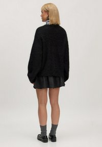Schwarzer, flauschiger Strickpullover mit überschnittenen Schultern, kombiniert mit einem schimmernden schwarzen, ausgehenden Rock. Graue Socken mit schwarzen Schuhen runden das Outfit ab.