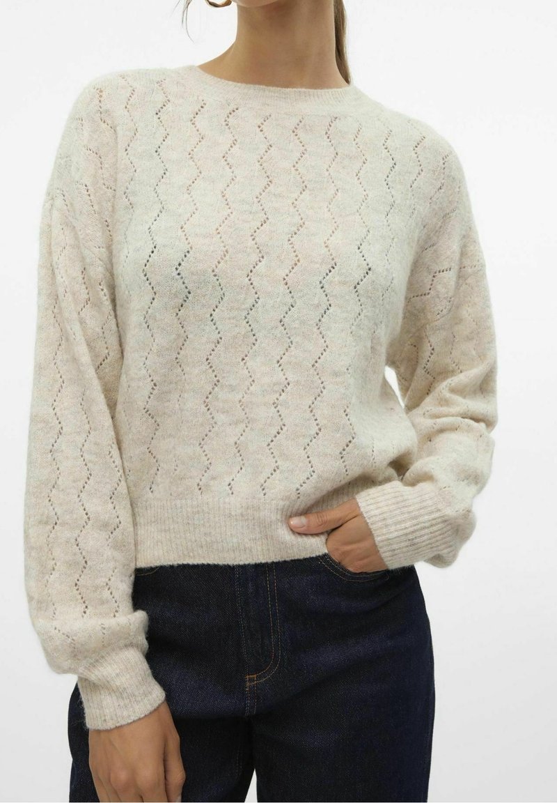 Pull en maille beige avec motif ajouré en zigzag et poignets côtelés, porté avec un jean bleu foncé, main placée dans la poche.