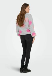 Femme aux longs cheveux bruns portant un pull gris à pois roses, un jean skinny noir et des bottines noires, se tenant de côté.