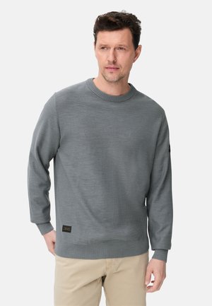 Homme portant un pull gris uni à manches longues et un pantalon beige, debout avec une main dans la poche, regardant légèrement sur le côté.