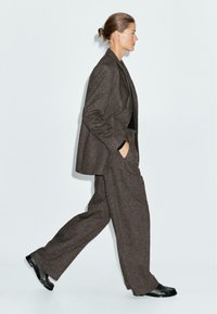 Donna che cammina verso sinistra, indossando un completo in lana marrone con pantaloni larghi, un blazer morbido, scarpe nere e i capelli raccolti in uno chignon.