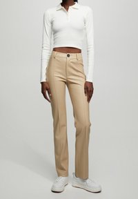 Polo blanc cintré à manches longues avec col, associé à un pantalon beige sur mesure avec un bouton et des coutures latérales. Porté avec des baskets blanches.