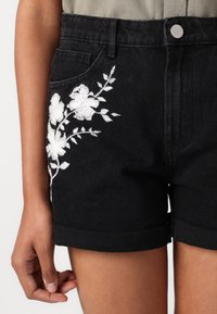 Svarte denimshorts med rullkanter, med hvit blomsterbroderi på venstre side og en frontlomme. Sølvfarge knappeutstyr synlig.