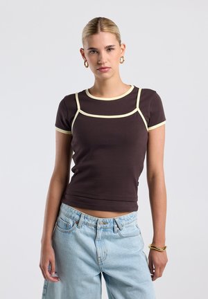 Jonge vrouw met blond haar draagt een bruin aansluitend T-shirt met beige bies en lichtblauwe hoge taille jeans, staand tegen een effen achtergrond.