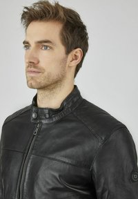 Schwarze Lederjacke mit Stehkragen, ausgestattet mit gesteppten Schulterakzenten und einem vorderen Reißverschluss. Glatte Textur und taillierte Passform.