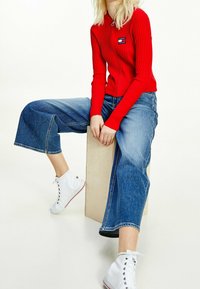 Rotes, geripptes Langarm-Polohemd mit kleinem Logo, kombiniert mit weiten blauen Jeansculotte und weißen Sneakers im High-Top-Stil.
