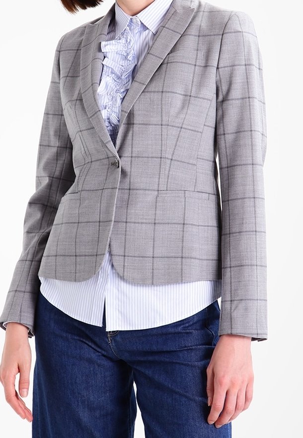 Femme portant un blazer à carreaux gris clair sur un chemisier rayé avec des volants, associé à un jean bleu foncé sur fond blanc.