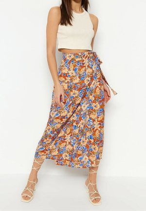 Femme portant un haut court sans manches blanc, une jupe midi portefeuille orange à fleurs, et des sandales à talons blanches à brides, se tenant devant un fond blanc.