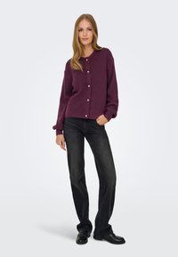 Bordeauxroter Strickcardigan mit rundem Ausschnitt, Knöpfen vorne und langen Ärmeln, kombiniert mit dunklen Jeans und schwarzen Schuhen.